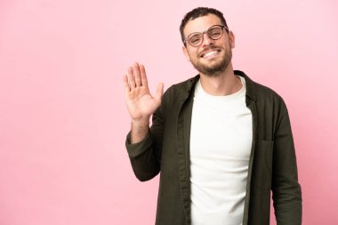 Pembe arka planda izole edilmiş genç Brezilyalı adam mutlu bir ifadeyle selamlıyor.