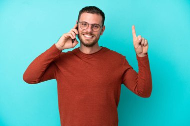 Mavi arka planda izole edilmiş cep telefonu kullanan genç Brezilyalı adam harika bir fikre işaret ediyor.