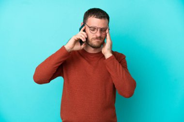 Mavi arka planda izole edilmiş ve baş ağrısı olan cep telefonu kullanan Brezilyalı genç bir adam.