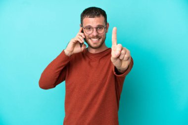 Mavi arka planda izole edilmiş cep telefonu kullanan genç Brezilyalı adam parmağını kaldırıyor ve gösteriyor.