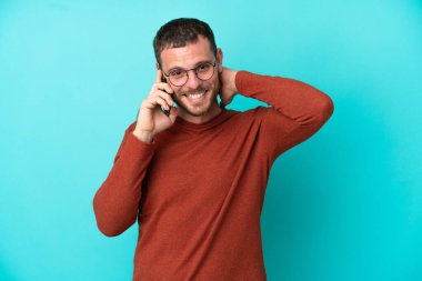 Mavi arka planda izole edilmiş cep telefonu kullanan genç Brezilyalı adam gülüyor.