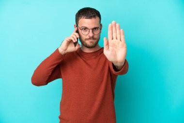 Mavi arka planda izole edilmiş cep telefonu kullanan genç Brezilyalı adam dur hareketi yapıyor