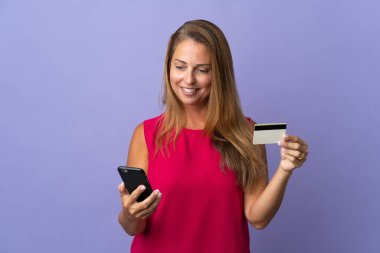 Mor arka planda izole edilmiş orta yaşlı Brezilyalı bir kadın cep telefonuyla kredi kartıyla alışveriş yapıyor.