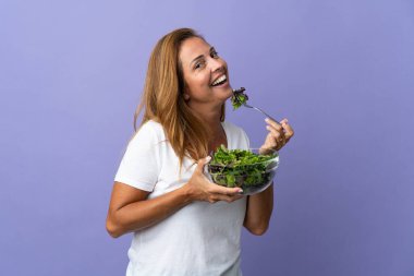Mor arka planda izole edilmiş orta yaşlı Brezilyalı bir kadın elinde mutlu bir ifadeyle bir kase salata tutuyor.
