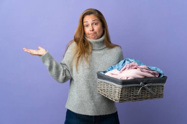 Orta yaşlı bir kadın mor arka planda izole edilmiş çamaşır sepetini tutarken ellerini kaldırırken şüpheye düşüyor.