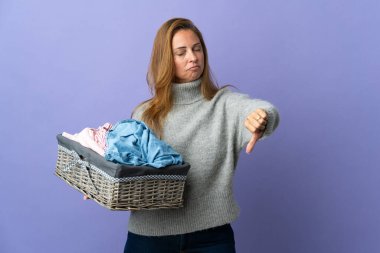 Mor arka planda izole edilmiş çamaşır sepetini tutan orta yaşlı bir kadın olumsuz bir ifadeyle baş parmağını gösteriyor.