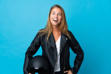 Mavi arka planda motosiklet kaskı tutan orta yaşlı bir kadın. Sürpriz bir yüz ifadesi var.