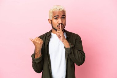 Pembe arka planda izole edilmiş genç Kolombiyalı yakışıklı bir adam. Yan tarafı işaret ediyor ve sessiz hareketler yapıyor.