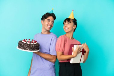 Genç melez çift ellerinde doğum günü pastası ve mavi arka planda izole edilmiş bir şekilde gülümseyerek omuzlarının üzerinden bakıyor.