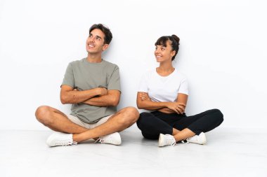 Genç melez çift, beyaz arka planda izole bir şekilde yerde oturuyor. Kollarını yan yana koyup gülümsüyorlar.