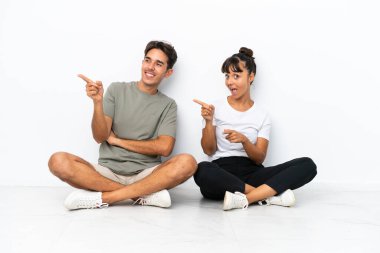 Yerde oturan genç melez çift beyaz arka planda izole edilmiş. Yan pozisyonda parmak gösteriyorlar.