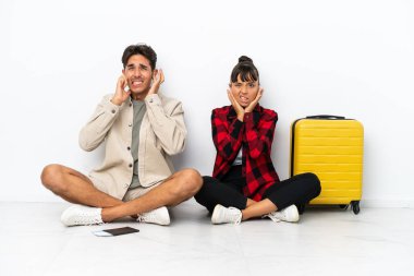 Genç melez ırk yolcuları, beyaz arka planda izole bir şekilde yerde oturuyorlar. İki kulağını da elleriyle kaplıyorlar.