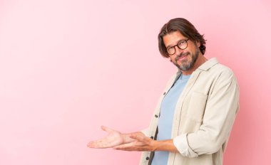 Kıdemli Hollandalı, pembe arka planda izole edilmiş. Davet etmek için ellerini yana uzatıyor.