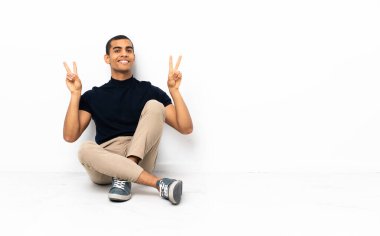 Yerde oturan Afro-Amerikan adam iki eliyle zafer işareti gösteriyor.