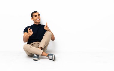 Afro-Amerikalı bir adam yerde oturuyor, ön tarafı gösteriyor ve gülümsüyor.