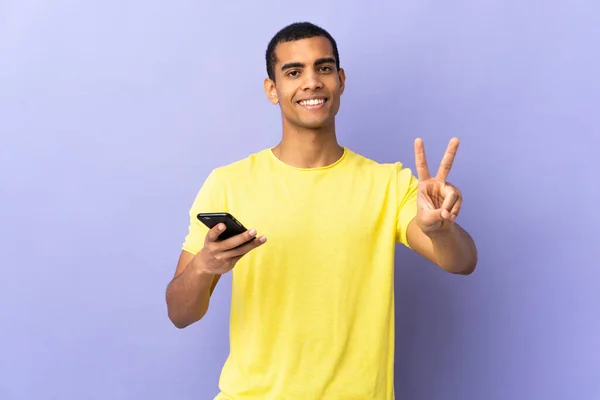 고립된 보라색 배경을 아프리카 미국인 핸드폰으로 웃으며 승리의 표시를 — 스톡 사진
