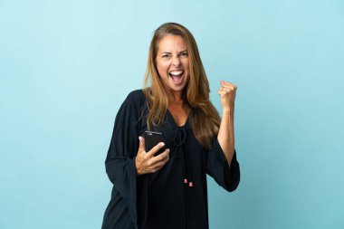 Orta yaşlı Brezilyalı kadın mavi arka planda izole edilmiş ve telefon zafer pozisyonunda.