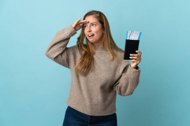 Seyahat eden orta yaşlı bir kadın mavi arka planda bir bavul tutuyor. Pasaport ve uçak biletleriyle uzakta bir yere bakıyor.