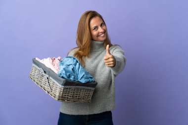 Orta yaşlı bir kadın mor arka planda izole edilmiş çamaşır sepetini tutuyor. Başparmakları havada çünkü iyi bir şey oldu.