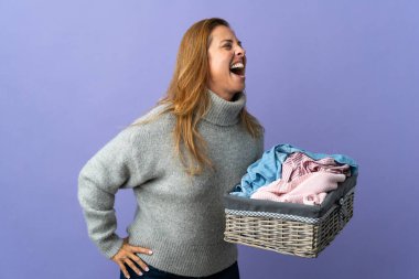 Orta yaşlı bir kadın mor arka planda izole edilmiş çamaşır sepeti tutuyor. Yan pozisyonda gülüyor.