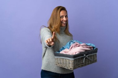 Mor arka planda izole edilmiş çamaşır sepetini tutan orta yaşlı kadın parmağını kaldırıyor ve gösteriyor.