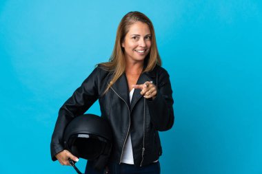 Mavi arka planda izole edilmiş motosiklet kaskı tutan orta yaşlı kadın şaşırmış ve ön tarafı işaret ediyor.