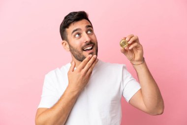 Elinde pembe arka planda izole edilmiş Bitcoin tutan genç adam gülümserken yukarı bakıyor.