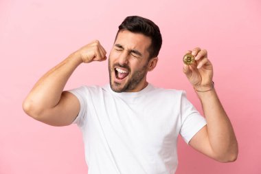 Elinde pembe arka planda izole edilmiş Bitcoin tutan genç adam güçlü bir jest yapıyor.