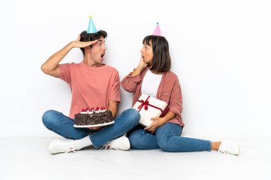 Genç melez çift, beyaz arka planda izole bir şekilde yerde oturmuş doğum gününü kutluyorlar. Şaşırmış ve şok olmuş bir yüz ifadesiyle.