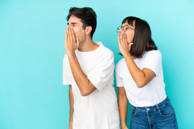 Genç melez çift mavi arka planda izole edilmiş ağızları sonuna kadar açık bağırıyorlar.