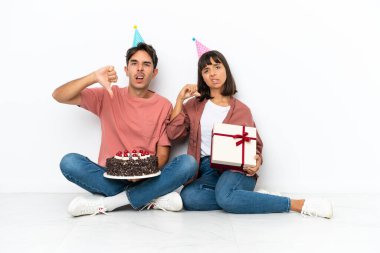 Genç melez çift, beyaz arka planda izole bir şekilde yerde oturmuş doğum gününü kutluyorlar.