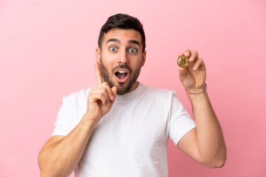 Elinde pembe arka planda izole edilmiş bir Bitcoin tutan genç adam çözümü bulmak için parmağını kaldırıyor.