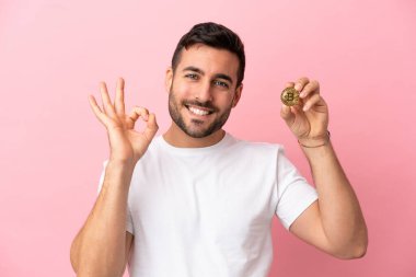 Elinde pembe arka planda izole edilmiş Bitcoin tutan genç adam parmaklarıyla tamam işareti gösteriyor.