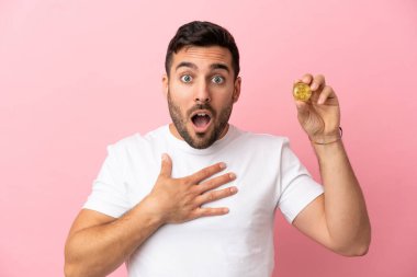 Elinde pembe arka planda izole edilmiş bir Bitcoin tutan genç adam sağa bakarken şaşırmış ve şok olmuştu.