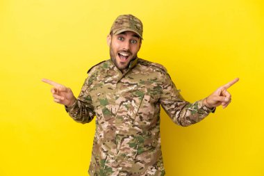 Sarı arka planda izole edilmiş asker parmak uçlarını işaret ediyor ve mutlu.