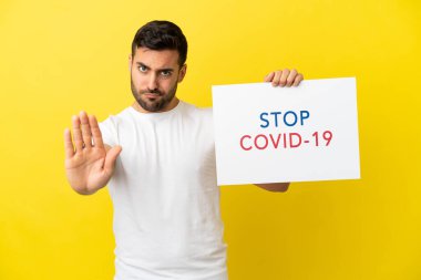 Sarı arka planda izole edilmiş yakışıklı beyaz bir adam elinde yazılı bir levhayla Covid 19 'u durdur ve dur işareti yapıyor.