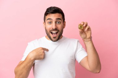 Elinde pembe arka planda, sürpriz bir yüz ifadesiyle izole edilmiş bir Bitcoin tutan genç adam.