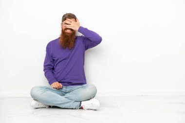 Beyaz arka planda izole bir şekilde yerde oturan beyaz tenli genç adam elleriyle gözlerini kapatıyor ve gülümsüyor.