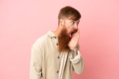 Pembe arka planda izole edilmiş genç beyaz adam yan tarafa bakarken sürpriz bir jest yaparak bir şeyler fısıldıyor.