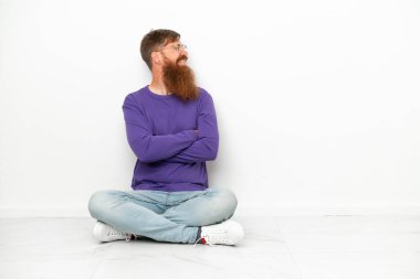 Beyaz arka planda soyutlanmış, mutlu ve gülümseyen genç, beyaz, kıpkırmızı bir adam yerde oturuyor.