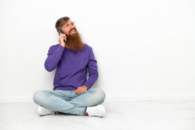 Beyaz arka planda izole bir şekilde yerde oturan Kafkasyalı genç adam cep telefonuyla sohbet ediyor.