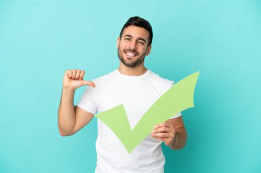 Mavi arka planda izole edilmiş yakışıklı beyaz bir adam elinde gururlu bir jestle bir çek simgesi tutuyor.