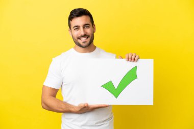 Sarı arka planda izole edilmiş yakışıklı beyaz bir adam. Elinde yeşil işaretli, mutlu ifadeli bir pankart var.