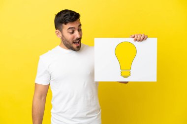 Sarı arka planda izole edilmiş yakışıklı beyaz adam elinde ampul simgesi olan bir pankart tutuyor.