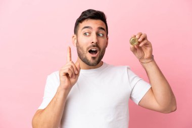 Elinde pembe arka planda izole edilmiş bir Bitcoin tutan genç adam bir fikri işaret ederek...