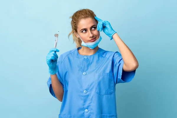 Młoda Kobieta Dentysta Trzyma Narzędzia Odizolowanym Niebieskim Tle Mając Wątpliwości — Zdjęcie stockowe