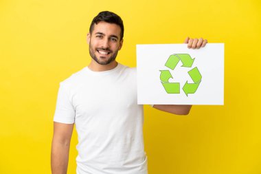 Sarı arka planda izole edilmiş yakışıklı beyaz bir adam elinde mutlu bir ifadeyle geri dönüşüm simgesiyle bir pankart tutuyor.