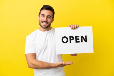 Sarı arka planda izole edilmiş yakışıklı beyaz bir adam. Elinde mutlu bir ifadeyle açık bir pankart tutuyor.