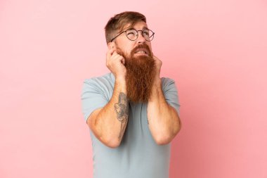 Pembe arka planda izole edilmiş genç beyaz adam hayal kırıklığına uğramış ve kulaklarını kapatmış.