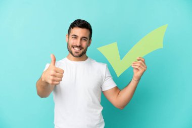 Mavi arka planda izole edilmiş yakışıklı beyaz bir adam elinde başparmağıyla bir kontrol simgesi tutuyor.